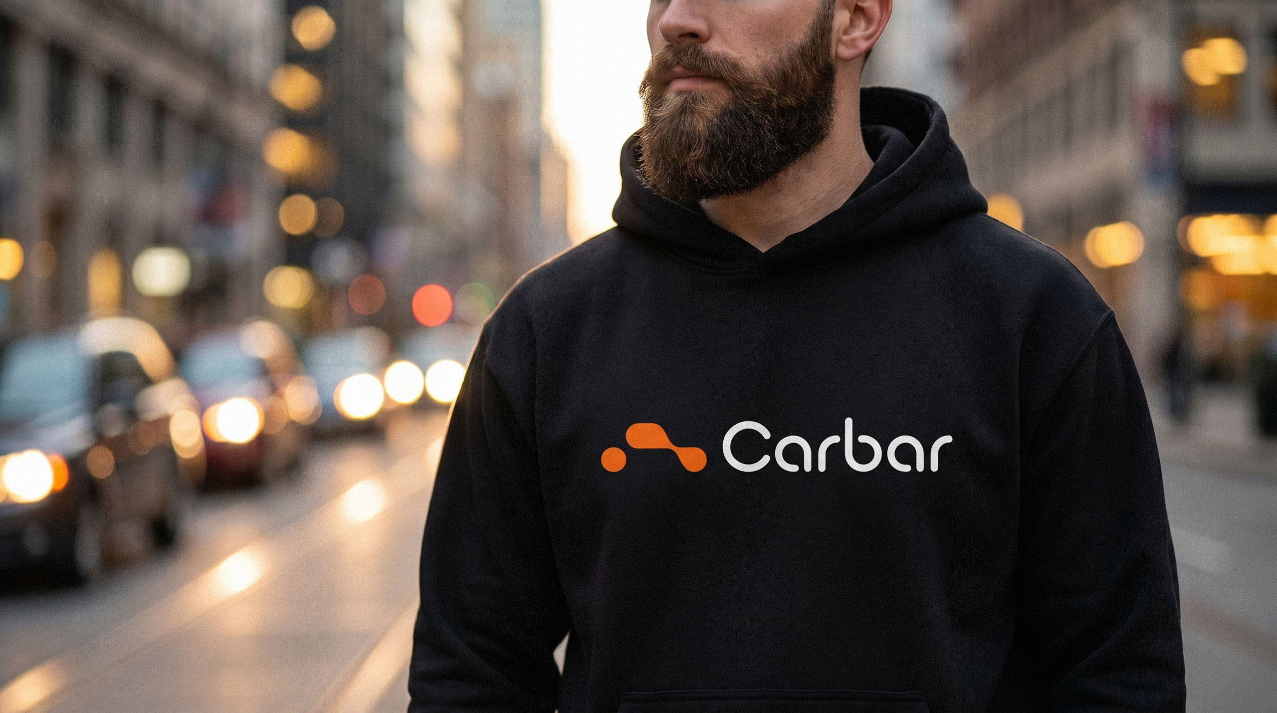 A bearded man wearing a black hoodie with the Carbar logo standing on a blurred city street - design by Brandforma Studio.