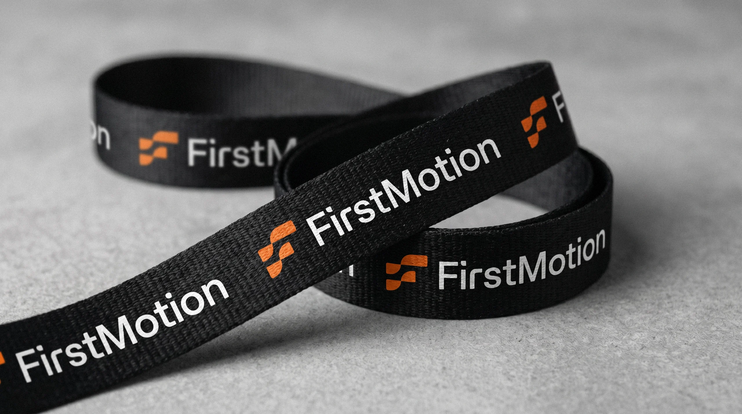 Black FirstMotion lanyard with repeated white logo coiled on a concrete surface
