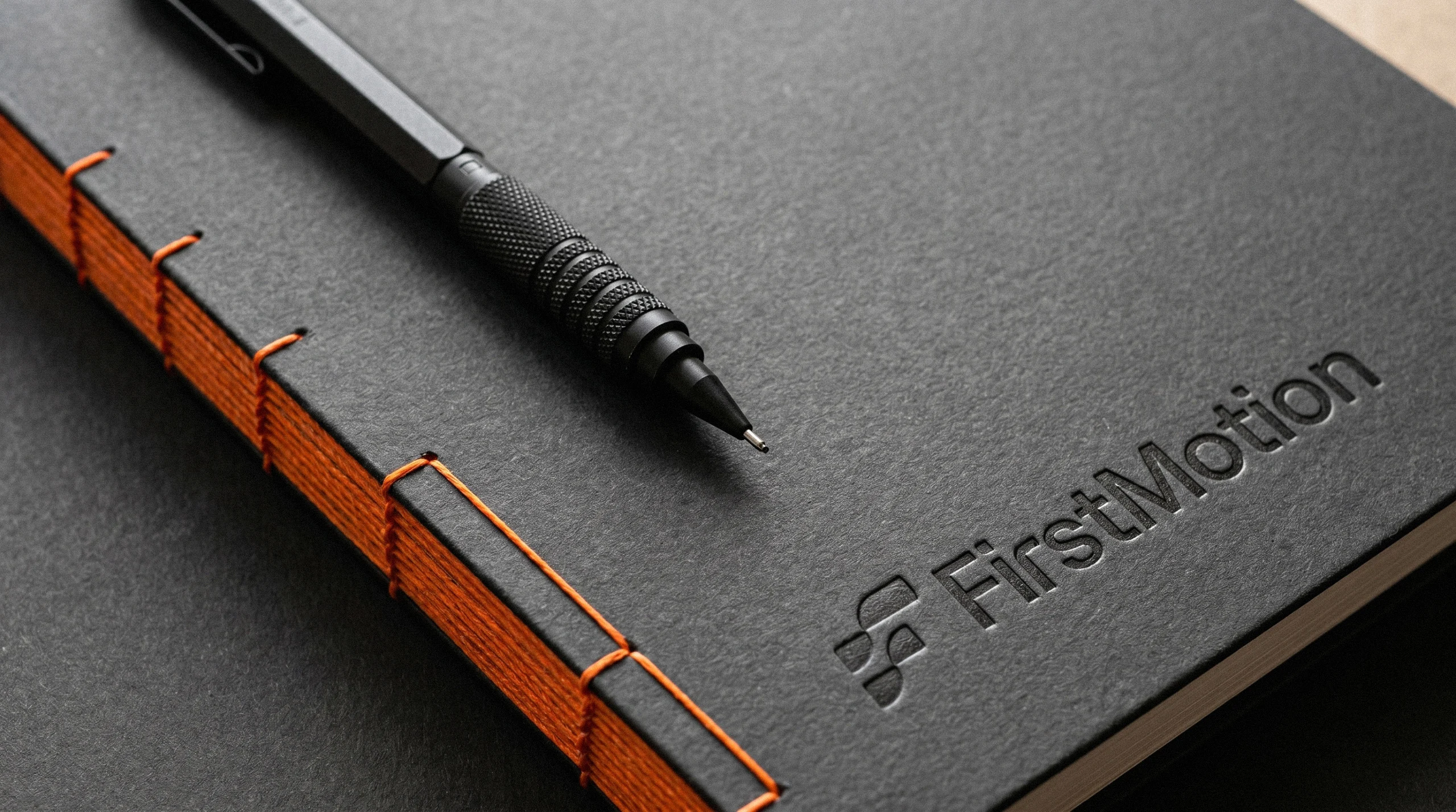 Dark FirstMotion notebook with orange binding and embossed logo, shown with a pen