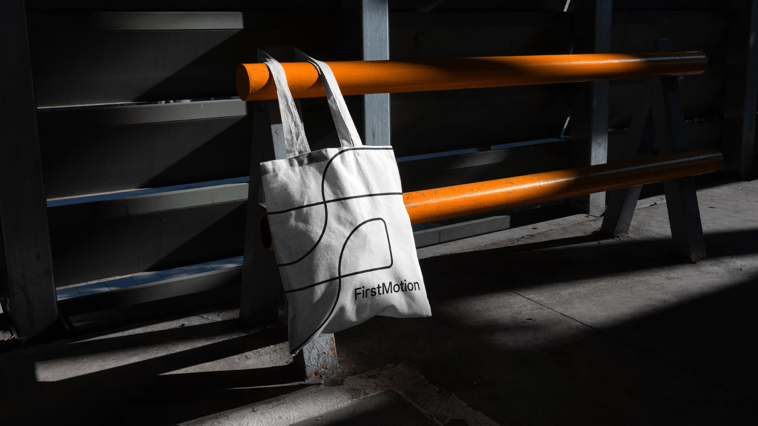 White FirstMotion tote bag with line-art mark hanging on industrial shelving lit by orange light
