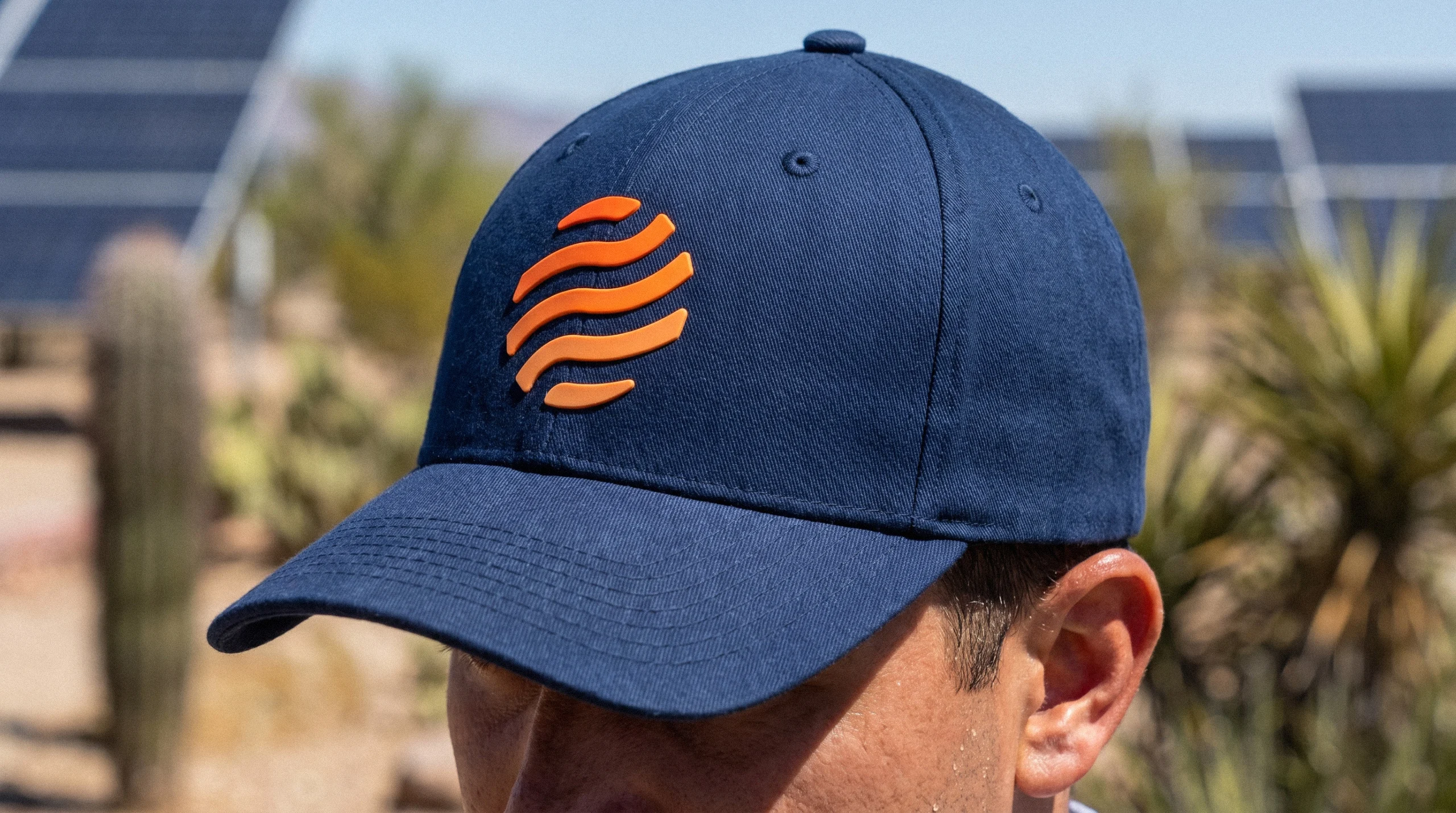 SolarPACES Branded Cap – Navy with Orange Mark Navy SolarPACES baseball cap embroidered with the orange solar mark, worn outdoors in front of solar panels